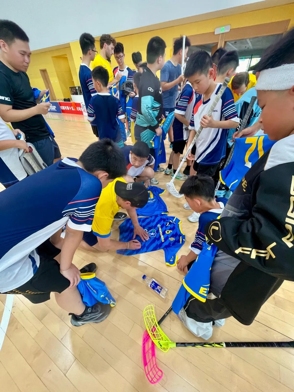 Young players gathered around a person signing blue and yellow Swedish team shirts on the floor of a sports hall