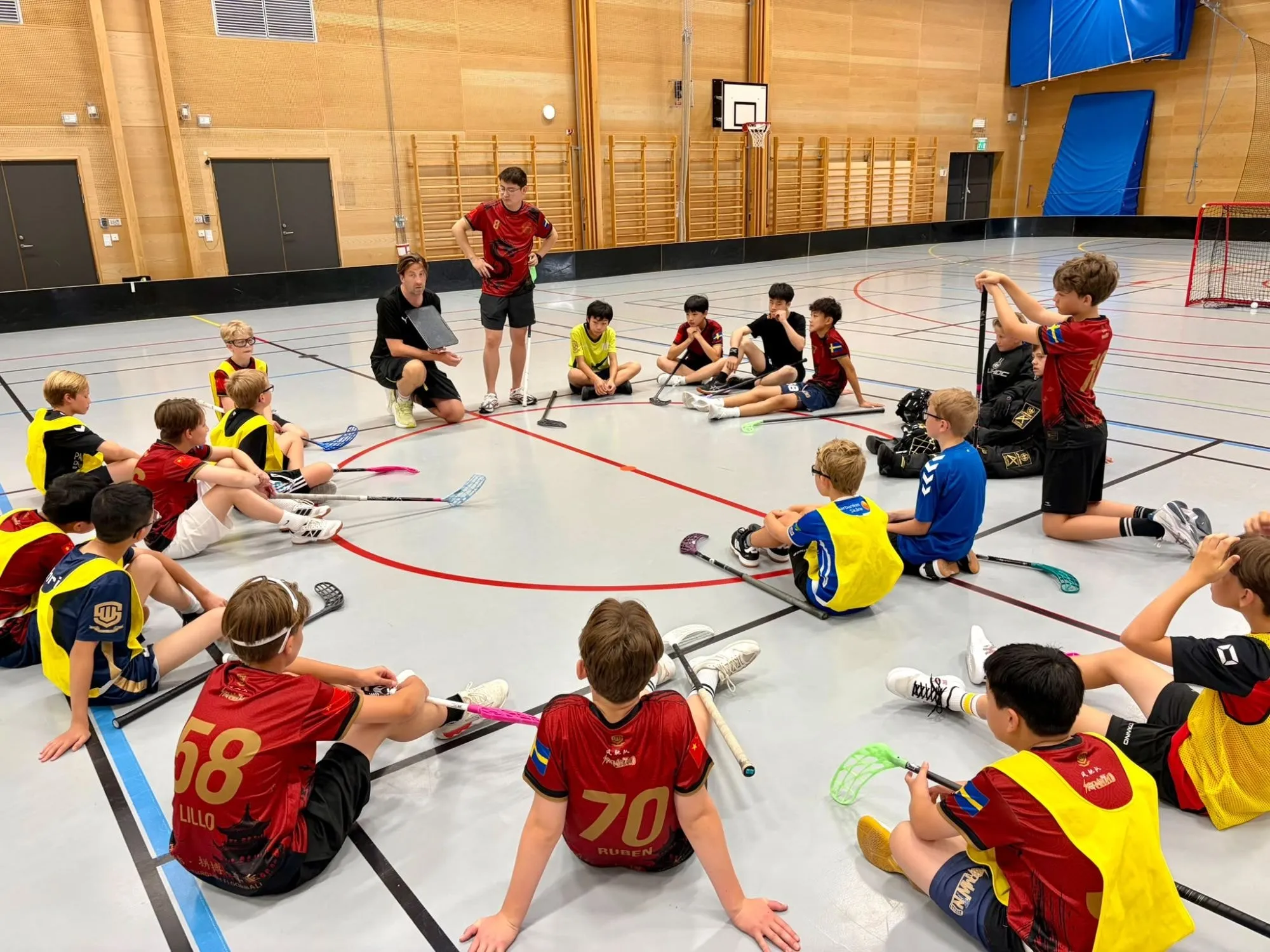 A coach with a tablet speaking to players sitting in a circle on the floor of a sports hall