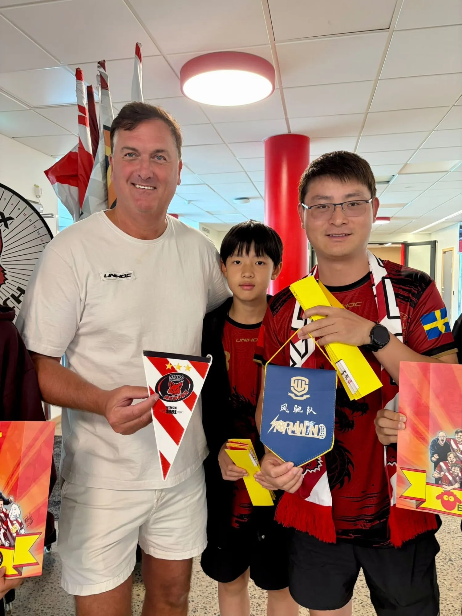 A coach and two players holding club pennants after a match, in red Floorball Asia jerseys