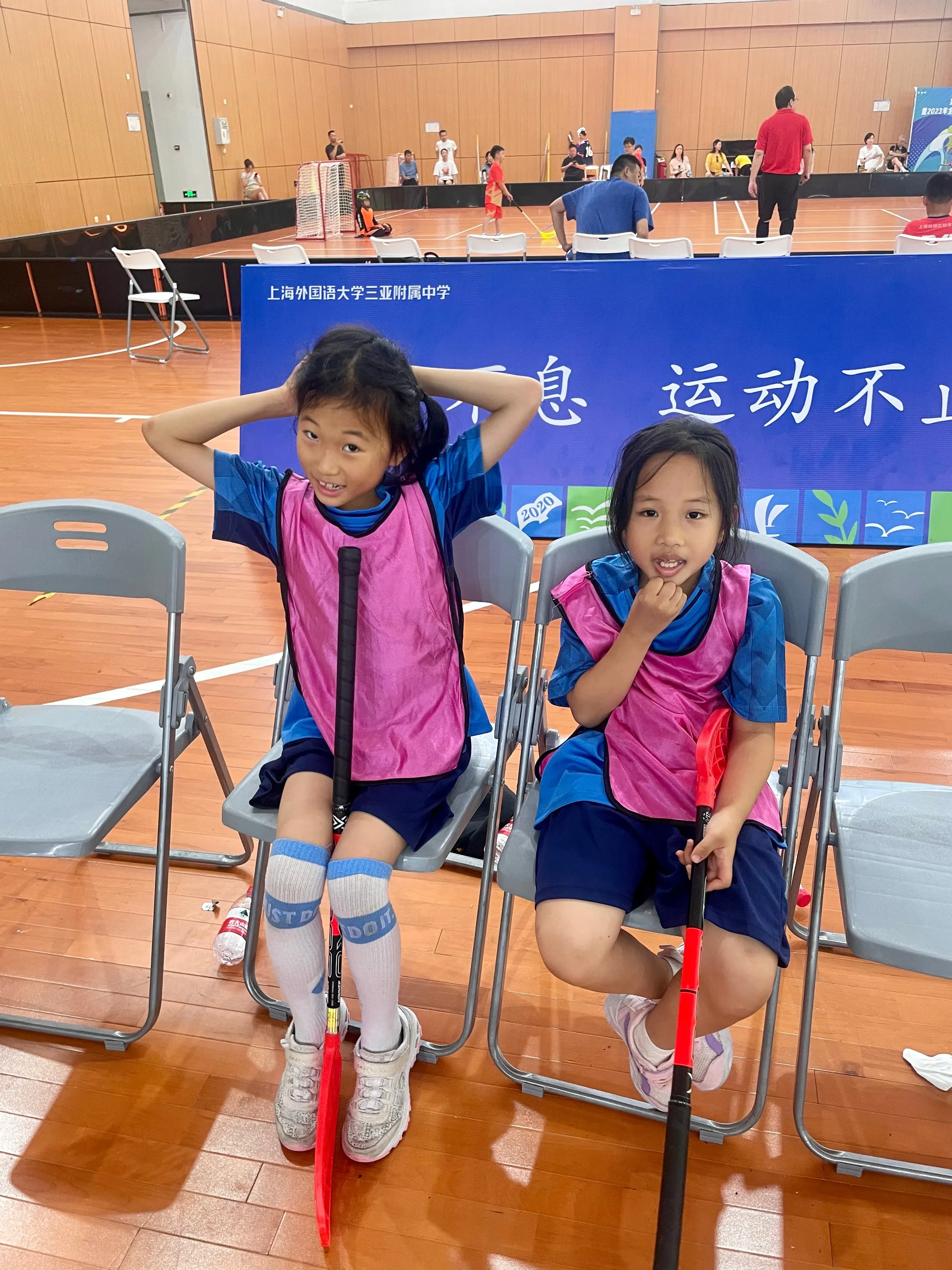 Two girls in pink bibs sitting on folding chairs with their sticks