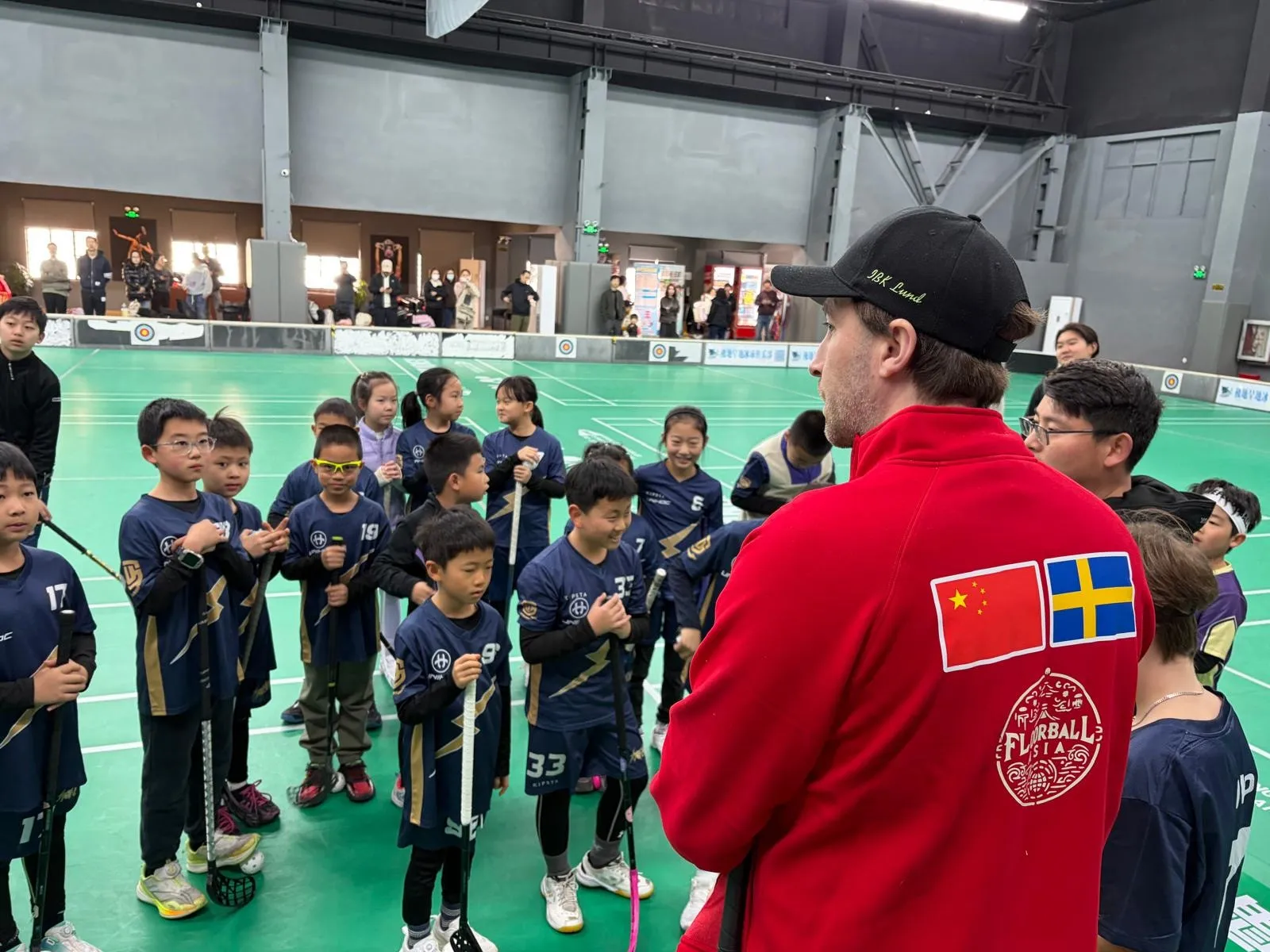 A coach in a Floorball Asia jacket talking to young players in navy jerseys on a green indoor court