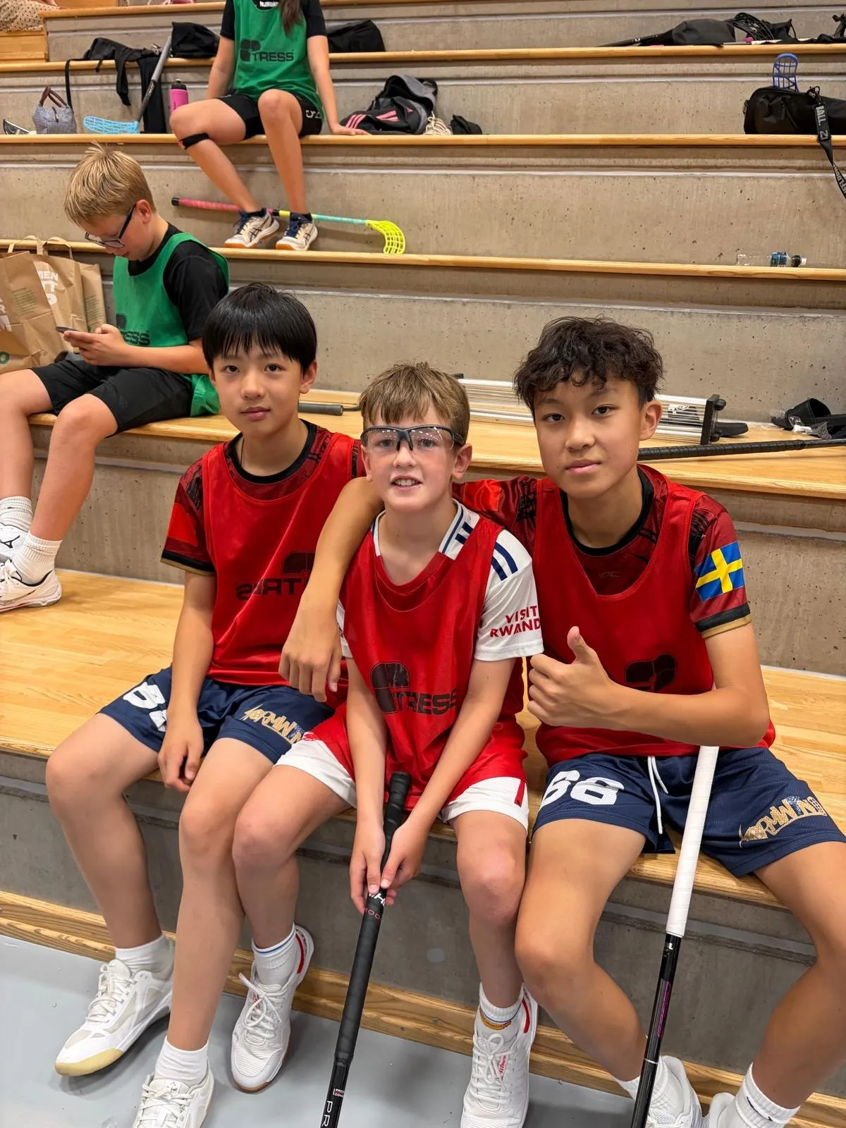 Three players in red bibs sitting together in the stands of a sports hall, one giving a thumbs up