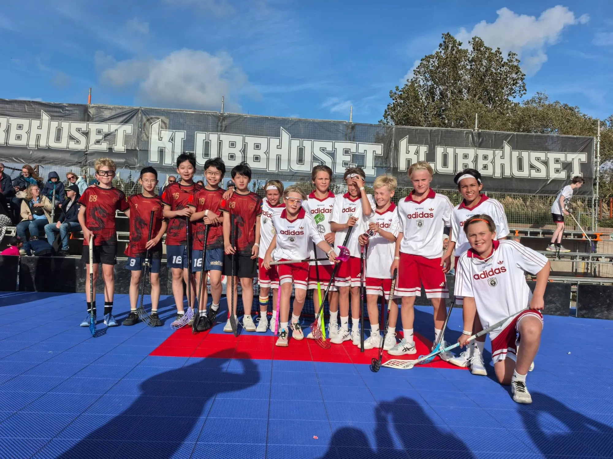 Floorball Asia players in red jerseys posing with opponents in white jerseys on a blue outdoor court